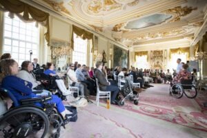 Grote groep mensen, sommigen in een rolstoel of met een geleidehond luisterend naar een spreker in een mooi versierde zaal.
