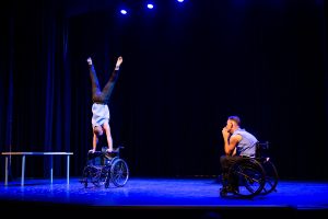 Een man zittend in een rolstoel en een man die een handstand maakt op een rolstoel